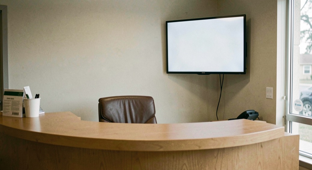 A reception desk in an imaging department with an empty chair behind it and a blank appointment screen mounted on the wall